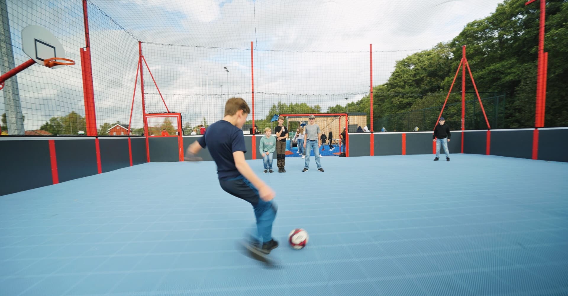 Flere gutter spiller fotball i en cageballbinge med røde stolper og basketkurver